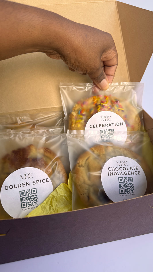 A hand placing an individually wrapped oversized bakery cookie into a Build Your Box gift box from Mrs Davids Creations.